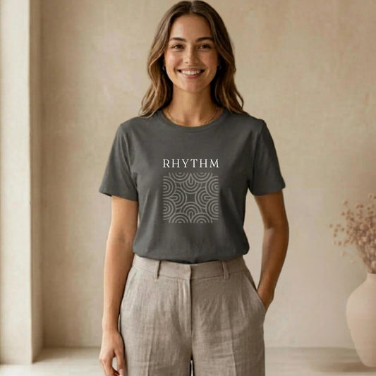 Woman wearing a gray t-shirt with 'RHYTHM' printed on it, standing in a room with a vase and plant.